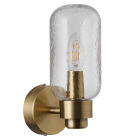 V�gglampan Tutan i m�ssing med cylindrisk, strukturerad glaskupa omsluter en synlig ljusk�lla och sprider ett varmt sken, monterad p� v�gg i n�rbild; en dekorativ och badrumsanpassad armatur fr�n Elbutik.se.