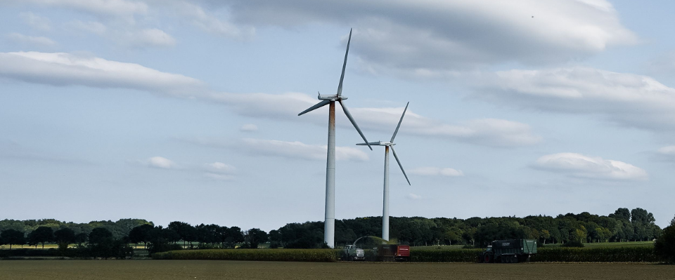 Tv&aring; vindkraftverk p&aring; &aring;kermark med tr&auml;d och molnig himmel i bakgrunden.
