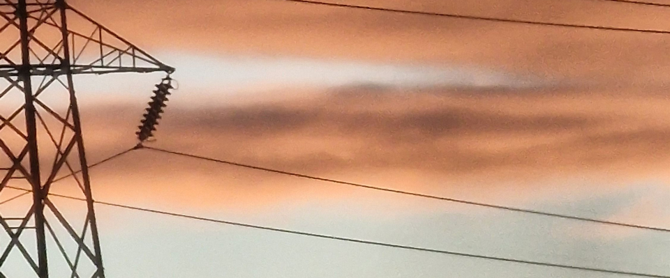 Del av en kraftledningsstolpe och elledningar mot en orangef&auml;rgad himmel.
