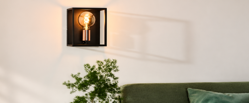 Svart fyrkantig v&auml;gglampa med synlig rund ljusk&auml;lla ovanf&ouml;r en gr&ouml;n soffa och en v&auml;xt.