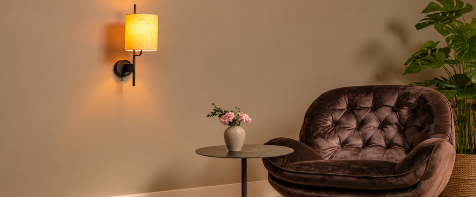 Svart v&auml;gglampa med cylindrisk ljus lampsk&auml;rm bredvid en brun f&aring;t&ouml;lj och ett runt sidobord.